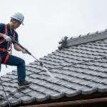 安全帯を装着した職人が、屋根の上で高圧洗浄を行っている。安全管理の徹底と丁寧な作業を示している