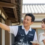 アップリメイクのユニフォームを着た専門家が、自宅の壁を指差しながら劣化状況を説明し、若い日本人夫婦が安心してその説明に耳を傾けている