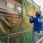 日本の専門業者がヘルメ-ットを着用し、メッシュシートで覆われた住宅で高圧洗浄を行っている作業風景