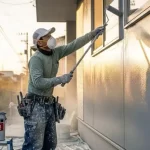 防臭マスクを着用した塗装職人が、外壁の中塗り（または上塗り）作業に集中している。臭いのピークとなる重要な工程での、プロフェッショナルな仕事ぶりを表している。