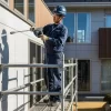 モダンな戸建て住宅の外壁塗装工事で、足場の上に乗った職人が高圧洗浄機を使い、壁を丁寧に洗浄している作業風景