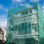 青空の下、外壁塗装工事のために足場と飛散防止ネットが組まれた都市部のモダンな20坪3階建て住宅