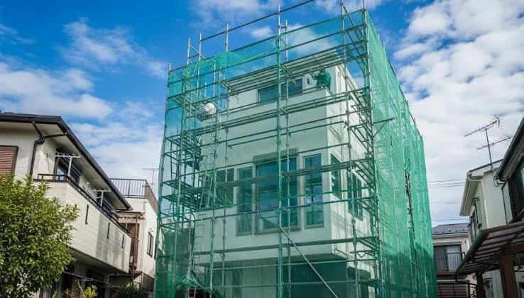 青空の下、外壁塗装工事のために足場と飛散防止ネットが組まれた都市部のモダンな20坪3階建て住宅