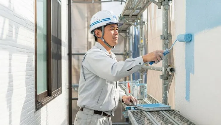 ヘルメットを着用した日本の塗装職人が、足場の上でローラーを使い、丁寧に外壁塗装を行っている様子