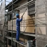 2階建てアパート全体に飛散防止のメッシュシートが張られた足場が組まれ、日本の塗装職人が外壁を高圧洗浄機で丁寧に洗浄している作業風景