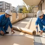 2人の日本の作業員が連携し、マンションの共用廊下で長尺シートの貼り付け作業を進めている様子